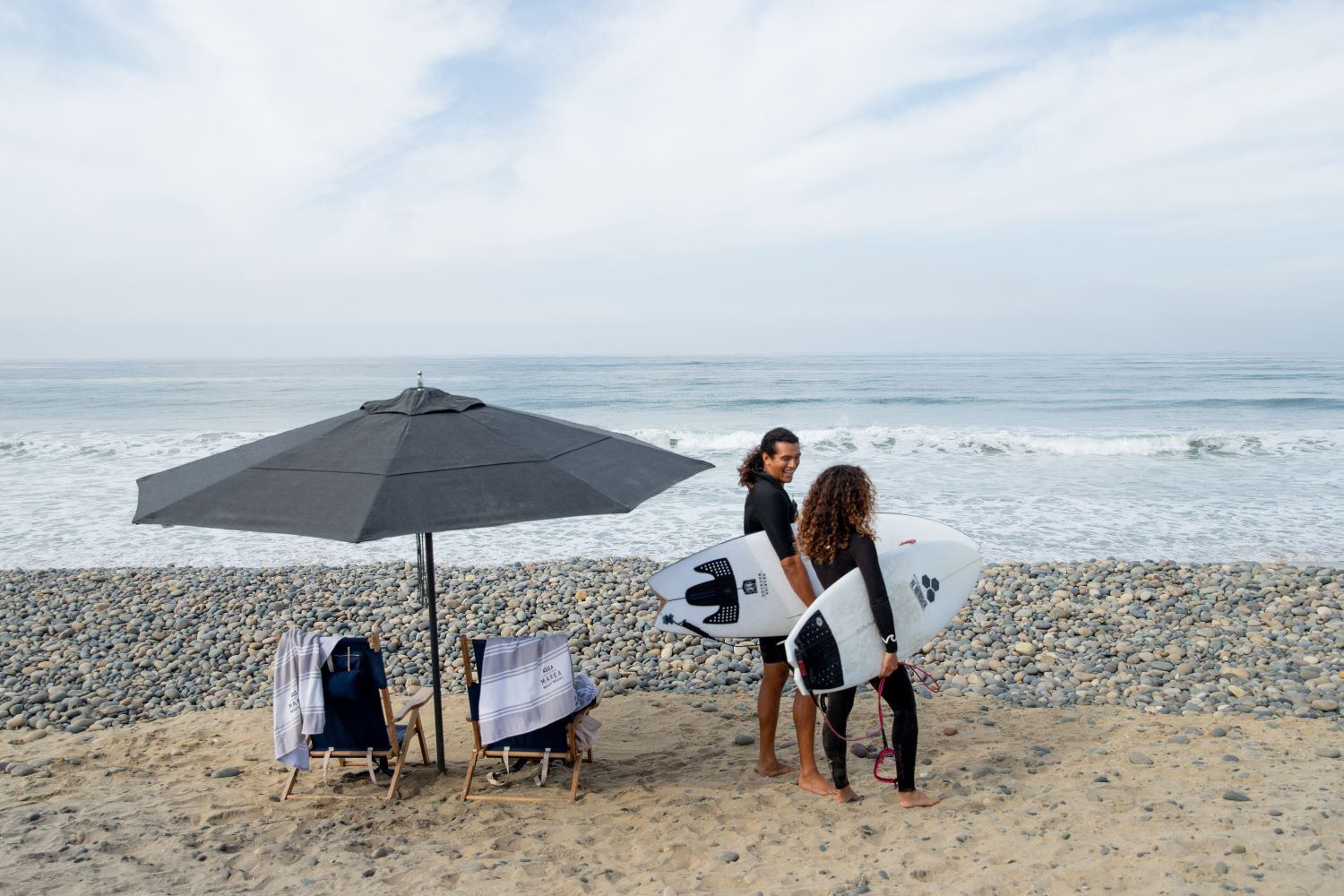 Two surfers carrying boards past a beach set-up with umbrella, chairs and resort towels on the cobblestone shore